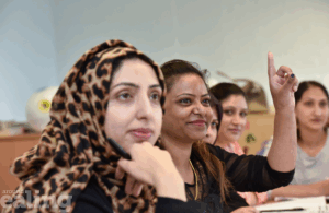 4 women sitting next to each other in what looks like a classroom. One woman has her hand raised