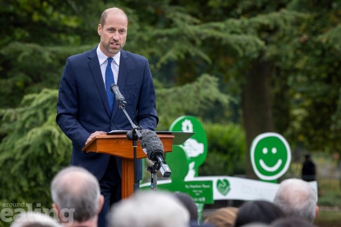 His Royal Highness, the Prince of Wales is standing at a lectern with a microphone giving a speech. He is wearing a navy blue suit and tie. There is part of the humanitarian assistance memorial behind him. He is in Gunnersbury Park.