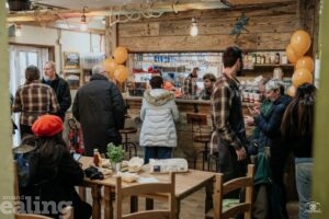 A cozy bistro interior with wooden decor, orange balloons, and shelves stocked with bottles and jars. Several people are gathered near the counter, some chatting and others ordering food. A table in the foreground is set with condiments, bread, and herbs, while a person in a red beret sits nearby. The atmosphere is lively and welcoming, reflecting a community event or opening celebration.