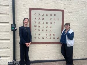 A photo of two women in front of an art installation piece. The art installation is orange brick square on white brick, containing orange text that reads "Orange are the bricks, white crush is the blossom".