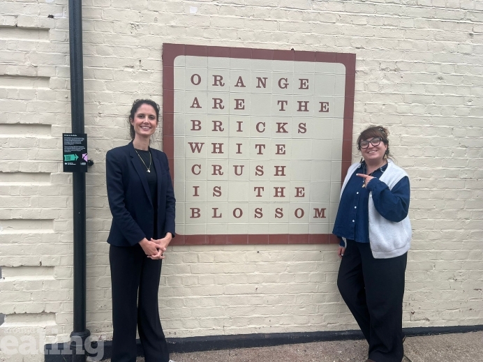 A photo of two women in front of an art installation piece. The art installation is orange brick square on white brick, containing orange text that reads "Orange are the bricks, white crush is the blossom".
