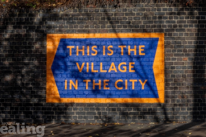 A photo of one of the installations. The art is a blue sign on an orange background, surrounded by black brick. The orange text on the sign reads "This is the village in the city".