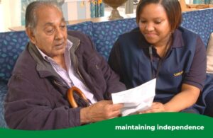 man looking at a form, sitting on a sofa, with a female carer leaning in to help him
