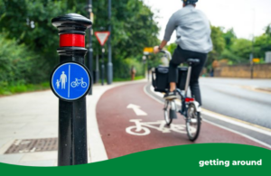 A person on a foldable bike wearing black trousers and a grey top with cycle helmet. They are cycling away from the camera along a smooth marked cycle lane. There is also a bollard with a cycle sign on it.