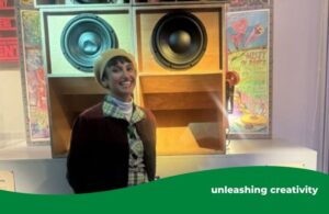 A smiling woman dressed in a plaid dress, light-colored boots, a beret and a dark jacket stands in front of a large vintage sound system with multiple exposed speakers and wooden enclosures. The display is part of an exhibit, with colorful posters and informational signs visible in the background, creating a vibrant and retro atmosphere.