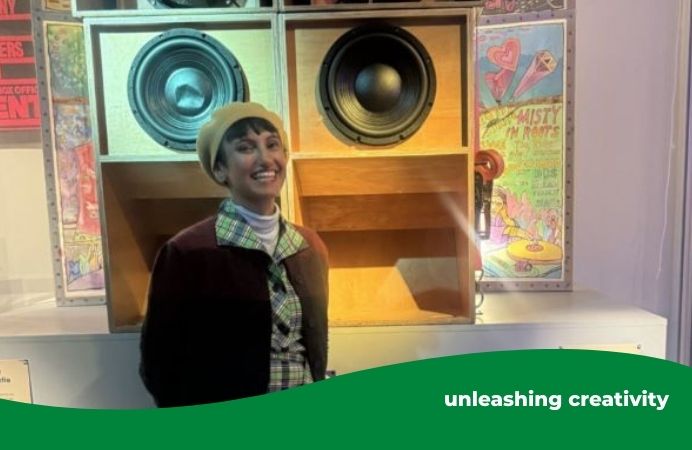 A smiling woman dressed in a plaid dress, light-colored boots, a beret and a dark jacket stands in front of a large vintage sound system with multiple exposed speakers and wooden enclosures. The display is part of an exhibit, with colorful posters and informational signs visible in the background, creating a vibrant and retro atmosphere.