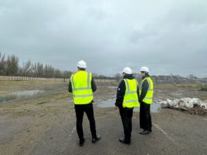 There are three people with their backs to the camera standing in a muddy building site with puddles in the background. They are all wearing white building site hard hats and flourescent yellow waistcoats.