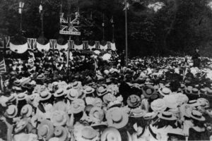 People gathered in a park for a celebration, with lots of them wearing straw hats and Union Jack flags and bunting flying on a stage in front of them