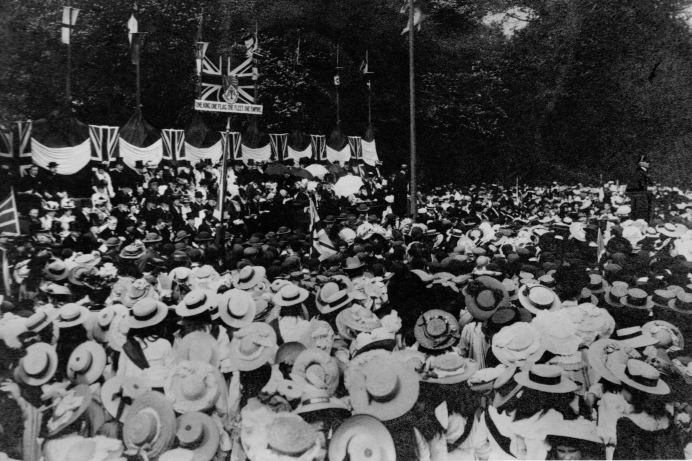 People gathered in a park for a celebration, with lots of them wearing straw hats and Union Jack flags and bunting flying on a stage in front of them