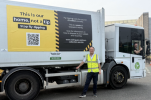 Councillor Paul Driscoll stood in front of a fly-tip branded refuse vehicle, wearing a high-visibility jacket. A driver is leaning out of the window with his thumb up