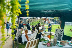 A group of people sitting outside under a green canopy painting on canvases.