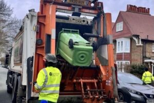 Person in a Greener Ealing branded high visibility jacket walking alongside a refuse vehicle with a green wheelie bin tipped into the back. Car and house in the background.