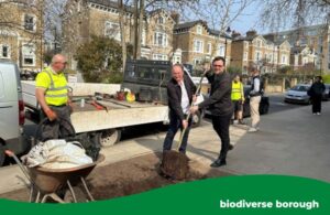 Councillors Paul Driscoll and Peter Mason are holding a tree which is about to be planted in the ground in front of them. There is worker with a hi-vis jacket on to help them and equipment including a wheelbarrow and spade.