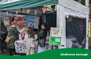 A community stall set up under a canopy at an outdoor event, displaying signs for the Ealing Repair Café and “Reduce, Reuse, Recycle.” Several people stand at the stall looking at clothing, textiles and repair demonstrations. The table is covered with patterned fabric, and banners about sustainability and local initiatives hang in the background. A green banner at the bottom reads “biodiverse borough.”