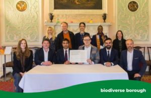 A group of 10 people sitting and standing behind a table with a white table cloth on in a lavish stately home. There are six people at the front seated at the table in a row. The centre two people from Hounslow and Ealing Councils are both holding the partnership agreement.