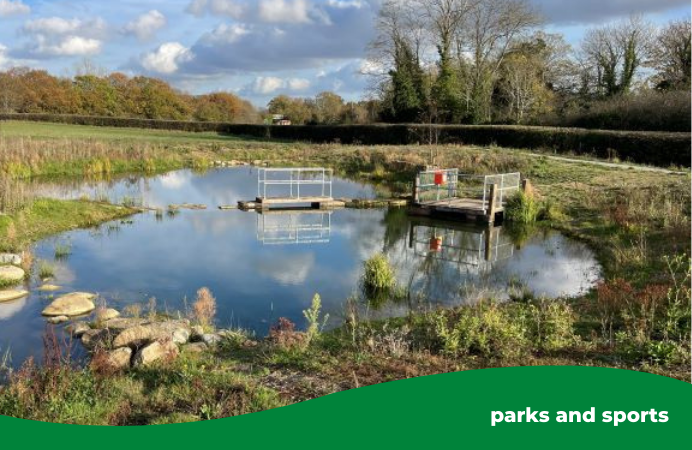 A pond in Glade Lane Canalside Park on a sunny day. It is surrounded by grass and there are trees in the background.