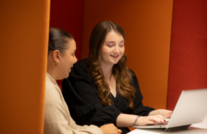 Two women sitting next to each other. One is typing into a laptop