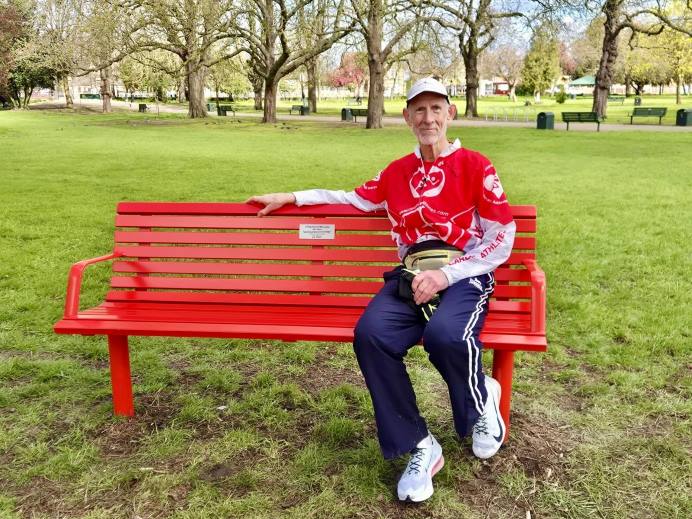 A man in a red t-shirt, blue jogging bottoms, wearing a baseball cap and white trainers is sitting on a bench.