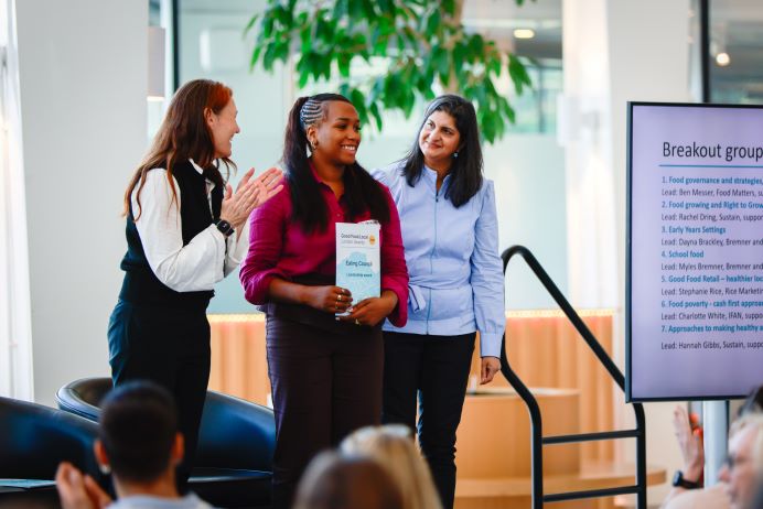 Three people stood on a stage. The person in the middle holding a certificate, the person on the left clapping, and the person on the right watching on.