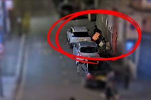 Blurred CCTV image with a red circle around a person wearing a high-visibility jacket dropping boxes on the pavement, next to a white car