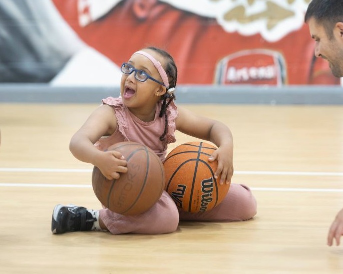 A little girl is wearing pink trousers and top is kneeling on the floor of a sports hall. She is holding 2 basketballs and is looking up to her right.