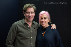 Two adults stand side by side against a dark background, facing the camera. One person wears a buttoned olive‑green shirt. The other wears a dark top and has pink‑coloured hair, with a small pair of orange scissors‑shaped jewellery pinned near the collar. One person’s arm is resting around the other’s shoulder.