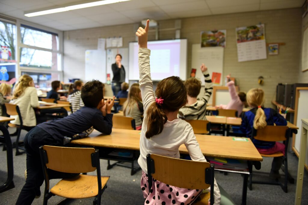 Classroom scene showing pupils at desks raising their hands as a teacher stands at the front.
