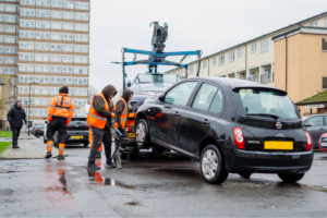 black nissan micra attached to a tow track, with 3 men in high visibility jackets and one plain-clothed man around it. Blocks of flats in the background