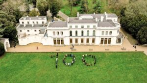 100 people are standing on the grass in front of Gunnersbury Park Museum to form the outline of the figure 100. This is to celebrate the park's 100th birthday.