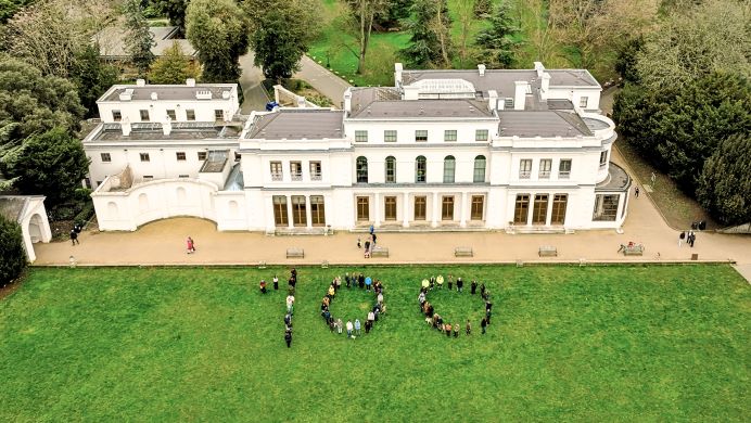 100 people are standing on the grass in front of Gunnersbury Park Museum to form the outline of the figure 100. This is to celebrate the park's 100th birthday.
