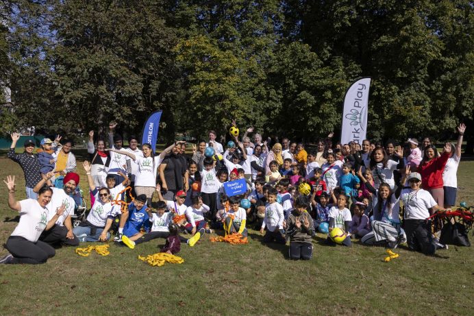 Large group of children and adults gathered in a park for a ParkPlay community sports session, raising hands and holding equipment. “Ready to play?” sign visible against a backdrop of trees on a sunny day