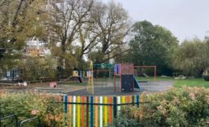 A playground with a climbing frame, slides, bench surrounded by colourful fencing and trees and bushes.