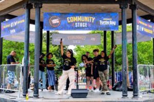 Performers in matching black T-shirts raise their arms on an outdoor stage under a “Community Stage” banner, with a microphone and speaker on the floor.