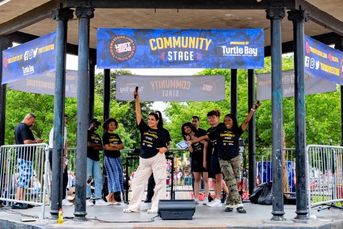 Performers in matching black T-shirts raise their arms on an outdoor stage under a “Community Stage” banner, with a microphone and speaker on the floor.