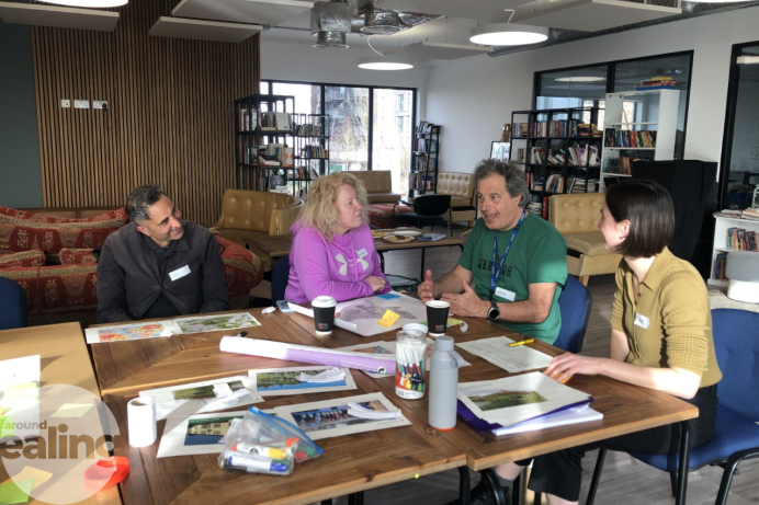 4 people sat around a table with various papers, pens and bottles on the table. Bookshelves and sofas are in the background