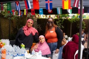 Two smiling women—one wearing a pink hijab and pink sunglasses, the other with black braided hair in an orange T-shirt and sunglasses—stand behind a child in a pink hijab making a peace sign. They are in a courtyard, in front of a table of refreshments under a canopy decorated with bunting.
