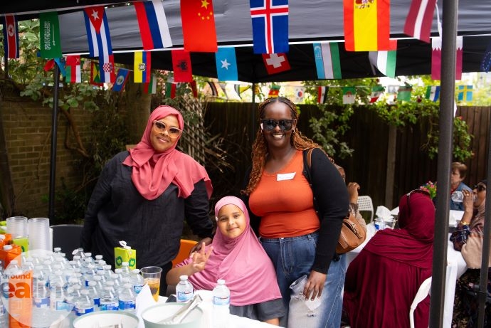 Two smiling women—one wearing a pink hijab and pink sunglasses, the other with black braided hair in an orange T-shirt and sunglasses—stand behind a child in a pink hijab making a peace sign. They are in a courtyard, in front of a table of refreshments under a canopy decorated with bunting.