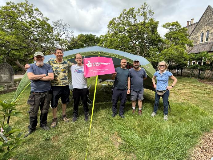 Volunteers from Ealing Parks Foundation standing on the grass at South Ealing Cemetery in front of a gazebo. There is a National Lottery sign on the gazebo.