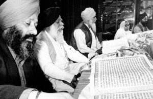 Three turban-wearing Sikh men with beards sitting reading a large manuscript. Other people are in the background