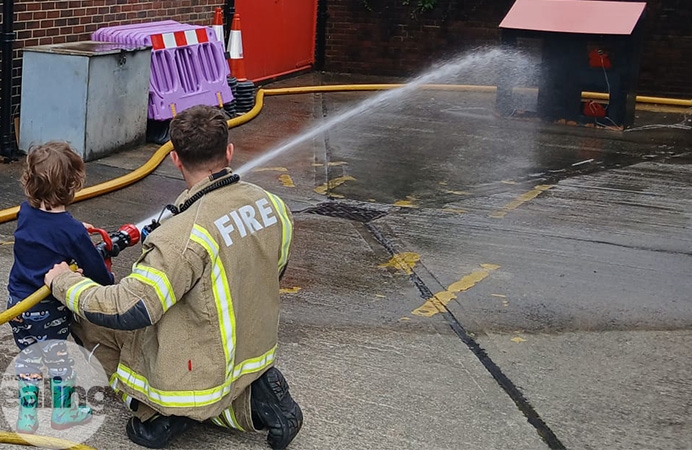 Fireman kneeling down, helping a child fire a jet of water from a firehose