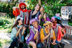 The cast of A Midsummer Night's Dream are grouped together. They are wearing bright costumes and some have flowers in their hair and glasses. They have bright coloured strips of fabric hanging around their necks and they are smiling.