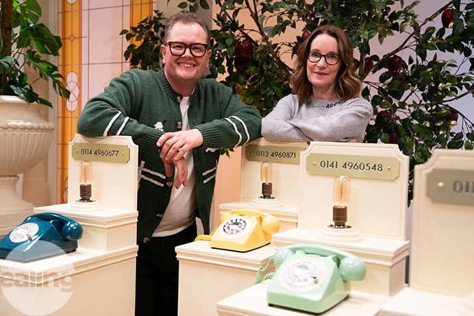 Man and woman standing next to several telephones. The man is Alan Carr and the woman is Susie Dent