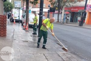 Man in high vid jacket using a wooden broom to sweep a pavement on a high street